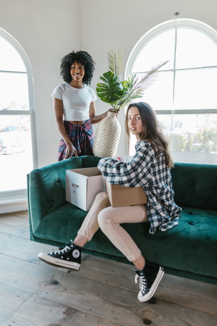 Two young women moving into a bright apartment, sitting on a green sofa with boxes.