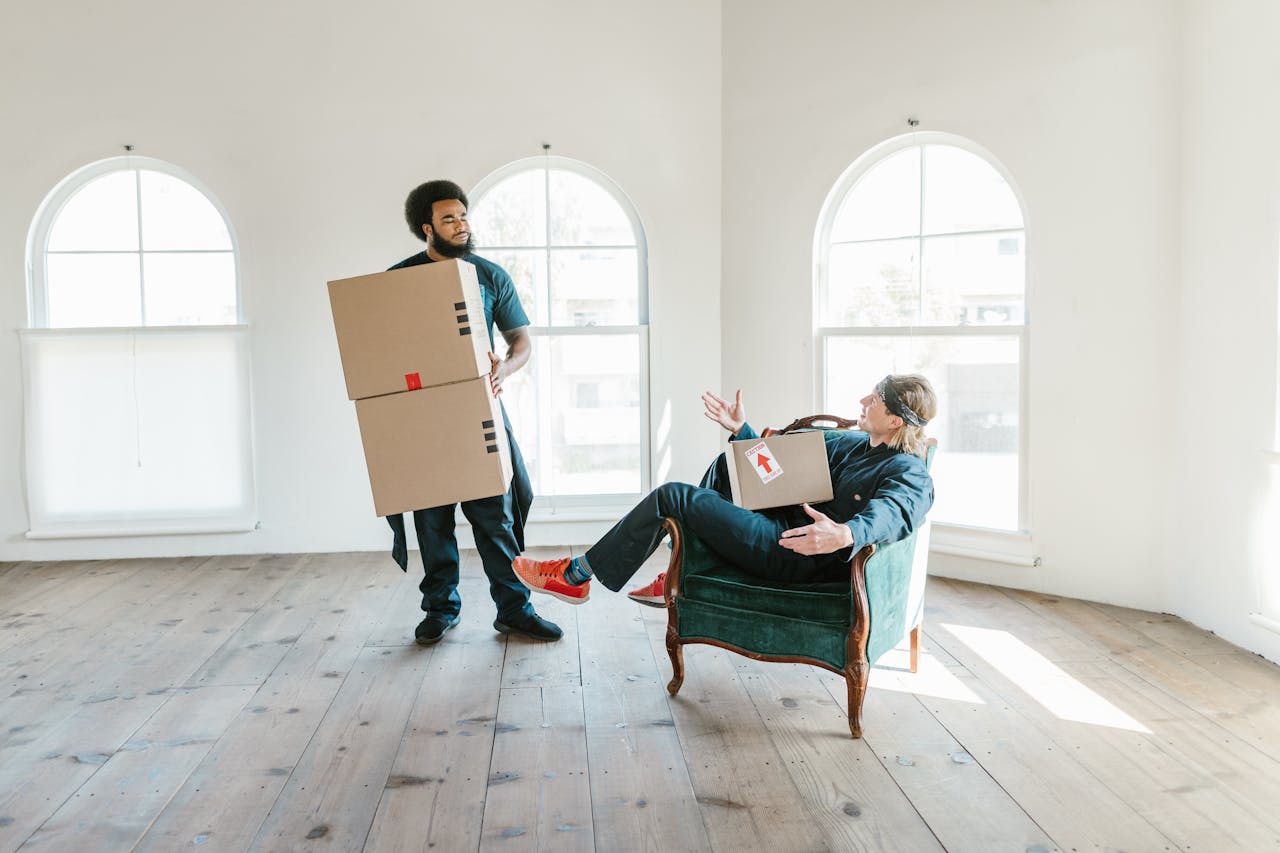 about-01 Two men, one sitting and one standing, moving boxes in a bright empty room with arched windows.